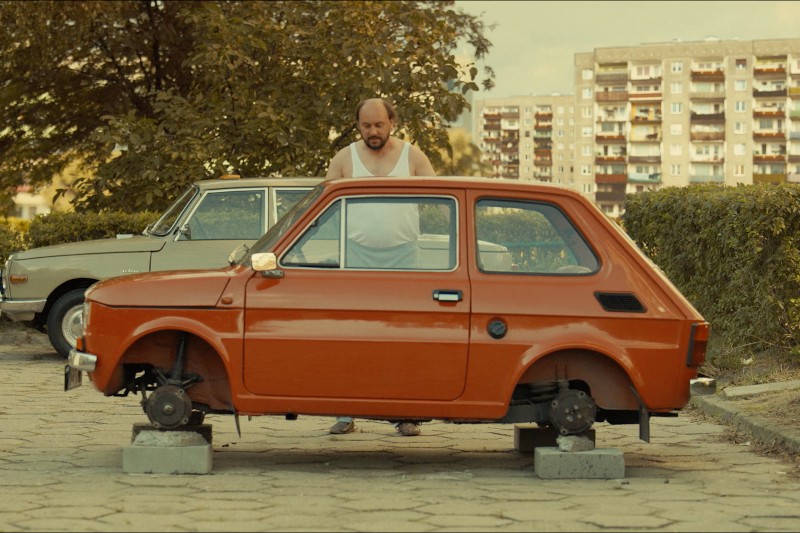 Mężczyzna w białym podkoszulku stoi za małym czerwonym samochodem, któremu brakuje kół i który stoi na pustakach na parkingu. W tle widać stary budynek mieszkalny i inny samochód.