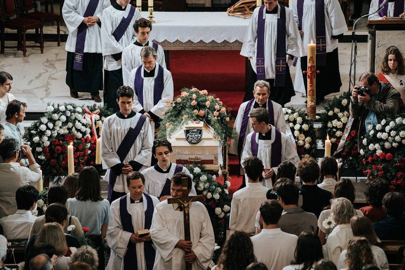 Grupa księży w białych i fioletowych szatach prowadzi nabożeństwo żałobne w kościele, otaczając trumnę ozdobioną kwiatami. Wokół zbierają się żałobnicy, a fotograf uwiecznia ten moment.