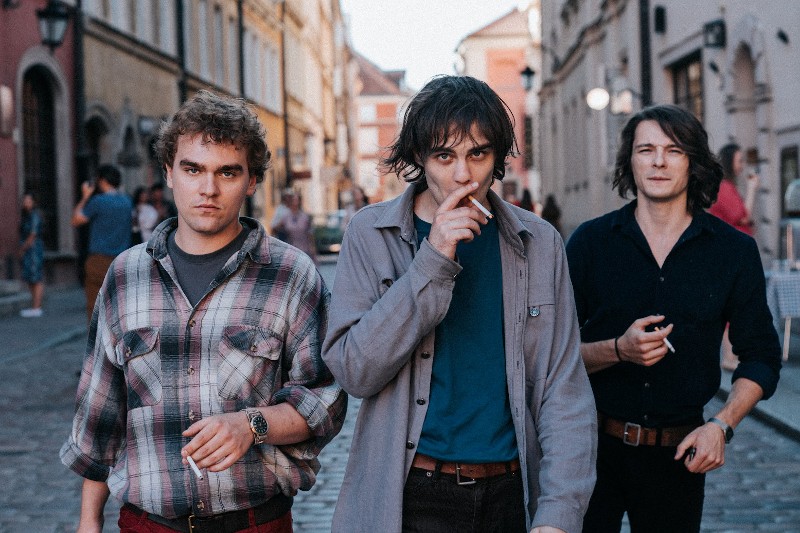 Trzech młodych mężczyzn idzie brukowaną ulicą w historycznym mieście. Dwóch ma papierosy, a wszyscy noszą luźne koszule. W tle widać stare budynki i spacerujących ludzi. Nastrój wydaje się poważny lub kontemplacyjny.