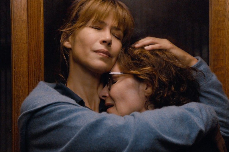 Dwie kobiety obejmują się blisko przy drewnianych drzwiach, z zamkniętymi oczami, wyrażając pocieszenie i wsparcie emocjonalne. Jedna ma krótkie brązowe włosy i okulary, druga ma jasnobrązowe włosy z grzywką.