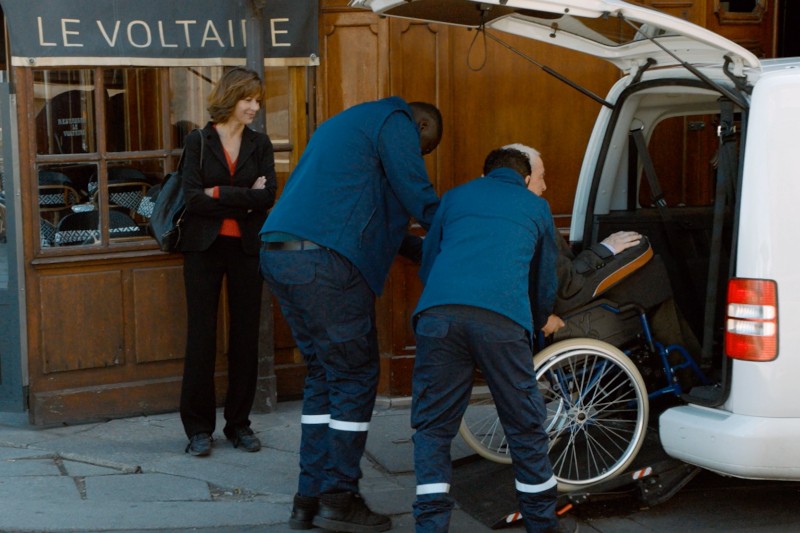 Dwóch ratowników medycznych pomaga osobie na wózku inwalidzkim wejść do białego vana przed budynkiem z napisem Le Voltaire, podczas gdy kobieta w czerwonej koszuli i czarnej kurtce stoi nieopodal i przygląda się.