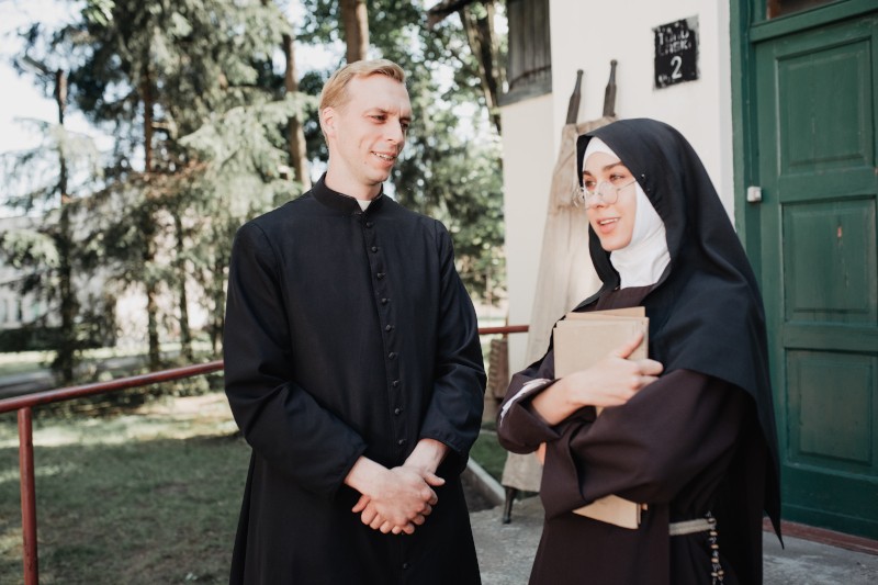 Ksiądz i zakonnica stoją przed budynkiem i rozmawiają. Zakonnica trzyma książkę i uśmiecha się, podczas gdy ksiądz słucha. W tle widać drzewa i zielone drzwi.