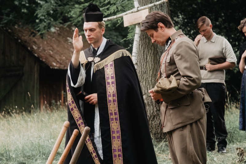 Ksiądz w czarnych szatach błogosławi coś na zewnątrz obok mężczyzny w mundurze wojskowym. Dwie inne osoby stoją za nimi, wszyscy na trawiastym, zalesionym terenie w pobliżu starego drewnianego budynku.