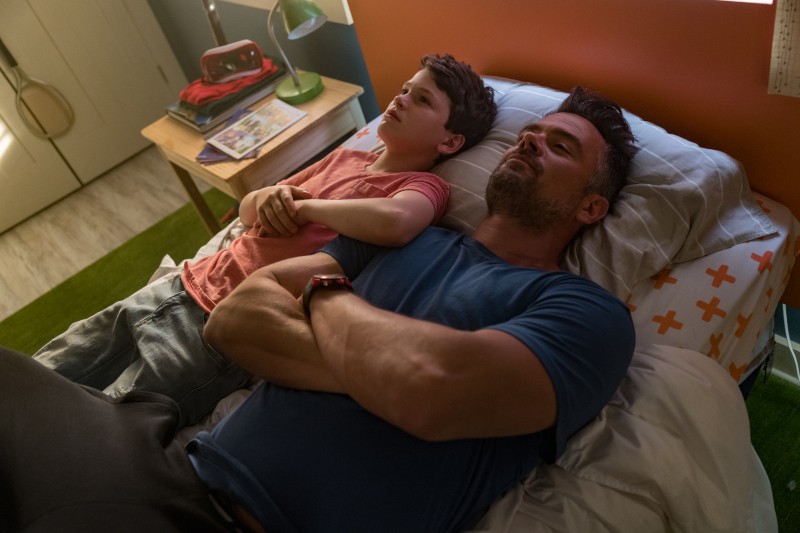 Mężczyzna i młody chłopiec leżą obok siebie na łóżku, wyglądając na zamyślonych. Chłopiec ma na sobie czerwoną koszulę, a mężczyzna niebieską. Obok łóżka stoi szafka nocna z książkami i lampą.