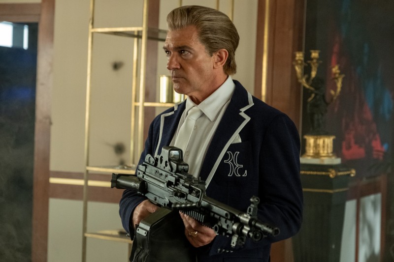 Mężczyzna z zaczesanymi do tyłu włosami, ubrany w granatowy blezer z białym wykończeniem, stoi w środku, trzymając duży czarny karabin, wygląda poważnie. W tle znajdują się złote półki i dekoracyjna rzeźba.