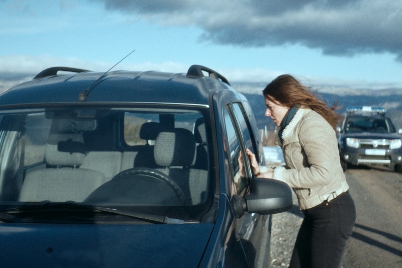 Kobieta o brązowych włosach w jasnej kurtce stoi obok zaparkowanego ciemnego SUV-a na wiejskiej drodze, zerkając niespokojnie przez okno; w tle widać inny pojazd pod zachmurzonym niebem.