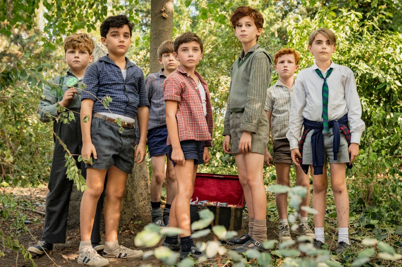 Siedmiu młodych chłopców stoi razem w zalesionym terenie, ubranych w szorty i koszule w stylu vintage. Wyglądają, jakby byli skupieni na czymś przed nimi, a za nimi na ziemi leży czerwone pudełko lub walizka.