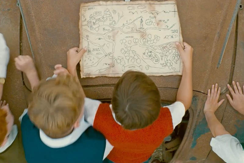 Trójka dzieci spogląda w dół na ręcznie narysowaną mapę skarbów, uważnie ją studiując na zardzewiałej metalowej powierzchni na zewnątrz. Ręce innych dzieci są widoczne na krawędziach, co sugeruje grupową aktywność lub przygodę.