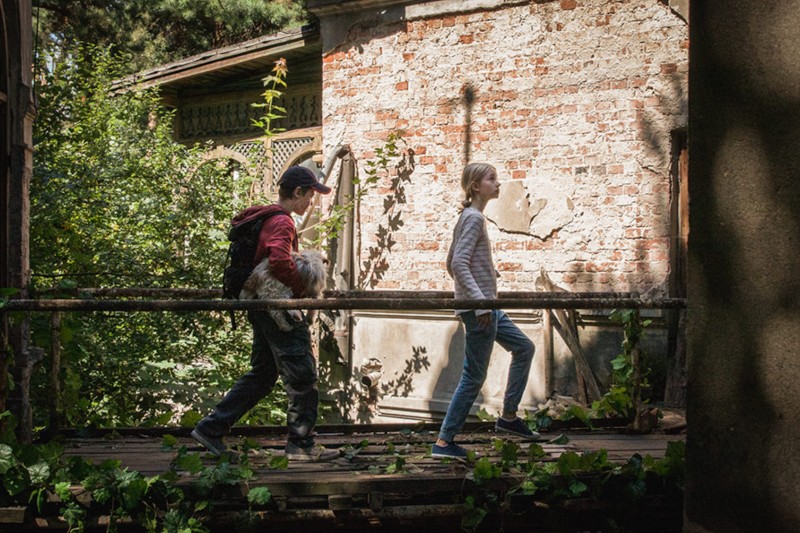 Dwie osoby przechodzą przez zniszczony drewniany most otoczony zielenią, obok starego budynku z cegły i kamienia. Jedna osoba niesie psa, podczas gdy światło słoneczne przebija się przez liście.