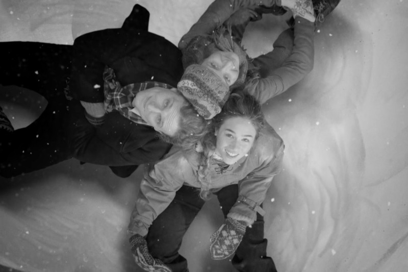 Trzy osoby leżą na plecach na śniegu, blisko siebie i uśmiechają się do kamery. Płatki śniegu spadają wokół nich, gdy noszą zimowe płaszcze, czapki i rękawiczki. Zdjęcie jest czarno-białe.