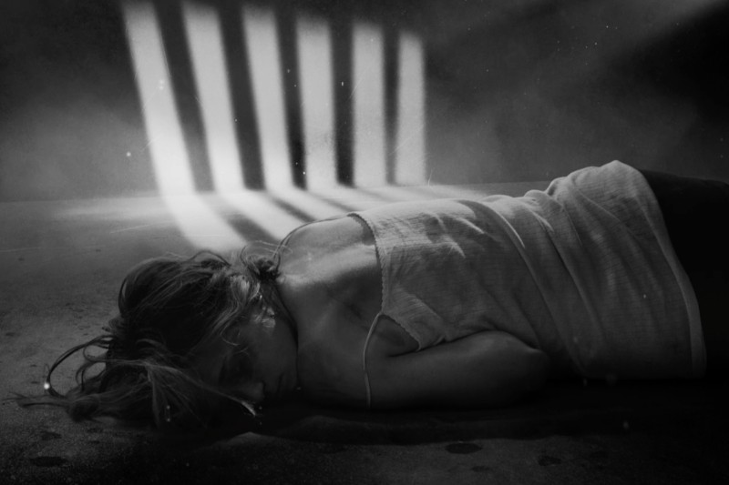 Kobieta leży na ziemi w słabo oświetlonym pokoju, oświetlonym światłem wpadającym przez zakratowane okna, rzucającym cienie sugerujące uwięzienie lub zamknięcie. Atmosfera jest ponura i napięta.