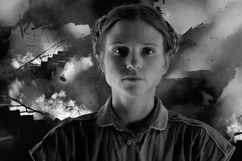 Kobieta z warkoczami patrzy poważnie przed siebie, ubrana w pasiastą koszulę. Za nią płomienie i dym unoszą się wśród ruin i połamanych schodów, tworząc dramatyczną, chaotyczną czarno-białą scenę.