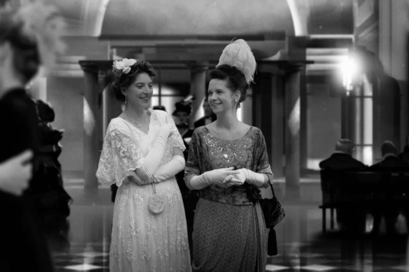Dwie kobiety w eleganckich, wczesno XX-wiecznych sukienkach i rękawiczkach uśmiechają się, idąc razem w wielkiej, ozdobnej sali. W tle widać łuki, kolumny i ludzi siedzących na ławkach. Obraz jest czarno-biały.