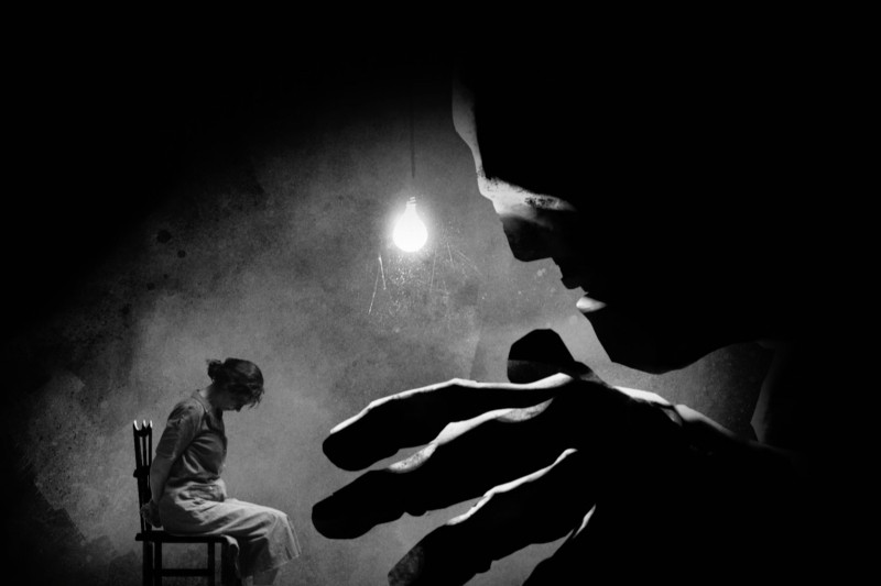 Kobieta siedzi na krześle pod wiszącą żarówką, pochylając głowę. Na pierwszym planie majaczy duża, złowroga, mroczna postać, której ręka wyciąga się w jej stronę w ciemnej, dramatycznej scenie.