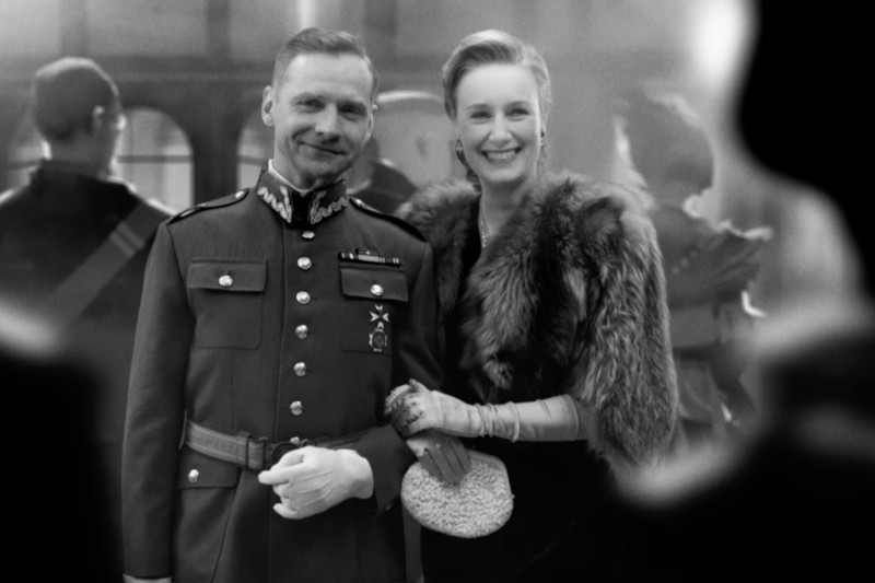 Uśmiechnięta para pozuje do zdjęcia; mężczyzna ma na sobie odznaczony mundur wojskowy, a kobieta olśniewającą sukienkę z futrzanym szalem, rękawiczkami i kopertówką. Scena wydaje się pochodzić z formalnego wydarzenia z przeszłości.