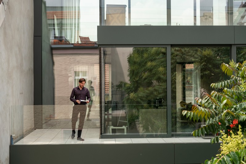 Mężczyzna stoi na nowoczesnym balkonie ze szklanymi balustradami, trzymając tablet. Za nim znajdują się duże szklane okna, w których odbijają się drzewa i pobliskie budynki. Na pierwszym planie widoczna jest zieleń.
