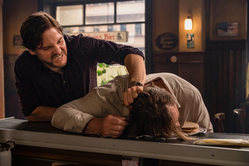 Mężczyzna agresywnie chwyta innego mężczyznę za włosy i uderza jego głową o ladę w słabo oświetlonej restauracji, obok której leży kanapka.