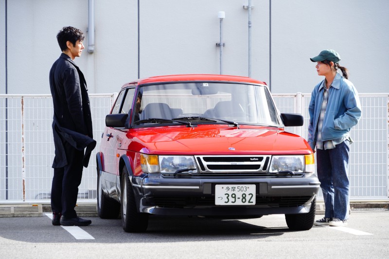 Dwie osoby stoją po przeciwnych stronach zaparkowanego czerwonego samochodu na parkingu, zwrócone do siebie twarzami. W tle widać biały płot i prosty budynek. Osoba po lewej stronie ma na sobie ciemne ubranie; ta po prawej nosi zieloną czapkę i swobodne ubranie.