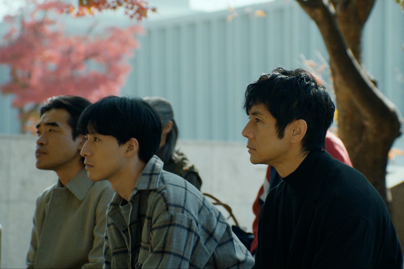 Trzech mężczyzn siedzi na zewnątrz, blisko siebie, uważnie patrząc przed siebie. W tle widać jesienne drzewa z czerwonymi liśćmi i nowoczesny budynek. Nastrój wydaje się spokojny i kontemplacyjny.