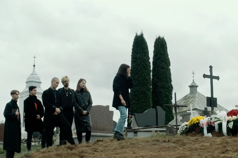 Grupa ludzi ubranych w ciemne ubrania stoi przy grobie na cmentarzu, niektórzy z pochylonymi głowami. Jedna osoba stoi osobno, zwrócona twarzą do grobu ozdobionego kwiatami, pod zachmurzonym niebem.