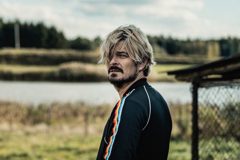 Mężczyzna z potarganymi blond włosami i brodą stoi na zewnątrz, w pobliżu jeziora, ubrany w czarną kurtkę z kolorowymi paskami na rękawach, patrząc przez ramię w stronę aparatu.