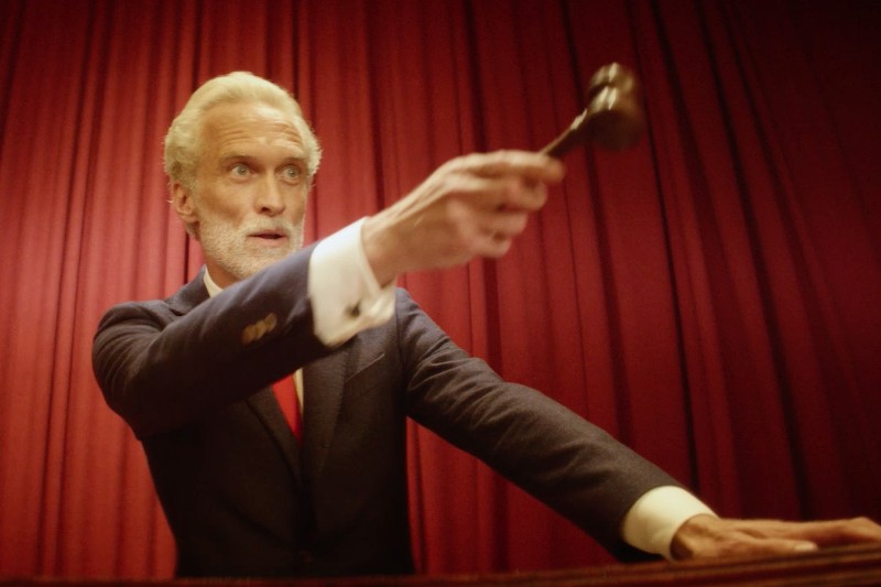 Starszy mężczyzna o białych włosach i brodzie, ubrany w ciemny garnitur i czerwony krawat, podnosi młotek, jakby zamierzał uderzyć, stojąc przed efektownymi czerwonymi zasłonami.