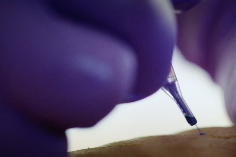 Zbliżenie dłoni w rękawiczce trzymającej maszynkę do tatuażu i nakładającej tusz na skórę. W miejscu kontaktu widoczne są igła i tusz.