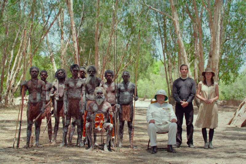 Grupa rdzennych mężczyzn z tradycyjnym malowaniem ciała i strojami stoi i siedzi na zewnątrz wśród drzew. Z przodu starszy rdzenny mężczyzna i biały mężczyzna w białym mundurze wojskowym siedzą na krzesłach. Mężczyzna w czerni i kobieta w sukience stoją obok nich.