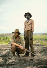 Mężczyzna w kapeluszu kuca z karabinem, a obok niego stoi drugi mężczyzna. Obaj ubrani są w solidne ubrania turystyczne, stoją na skalistym terenie z zielenią i odległymi wzgórzami w tle.