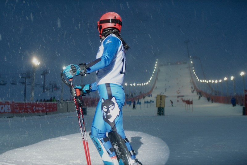 Narciarz ubrany w niebiesko-biały strój z wizerunkiem psa rasy husky na nodze, ubrany w czerwony kask, stoi nocą na zaśnieżonym stoku, trzymając kijki narciarskie, podczas gdy pada śnieg i światła oświetlają trasę narciarską przed nim.