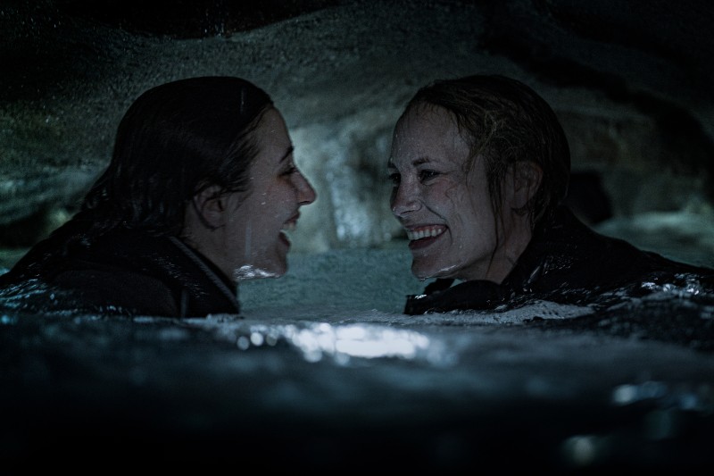 Dwie kobiety uśmiechają się do siebie, częściowo zanurzone w wodzie w ciemnej jaskini. Ich twarze są blisko siebie, mokre i oświetlone miękkim światłem, co tworzy poczucie połączenia i przygody.