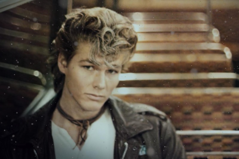 Młody mężczyzna z falowanymi blond włosami siedzi sam na wzorzystym siedzeniu w autobusie. Ma na sobie skórzaną kurtkę, białą koszulę i cienki szalik na szyi, patrząc zamyślony w kamerę.