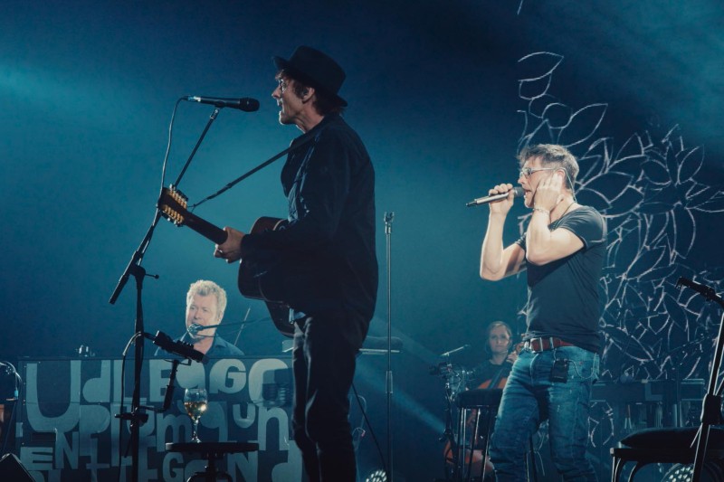 Na scenie występuje trzech muzyków: jeden gra na gitarze i śpiewa do mikrofonu, drugi śpiewa trzymając się za ucho, a trzeci siedzi przy pianinie. Scena jest oświetlona niebieskim światłem i ozdobiona artystycznymi wzorami tła.