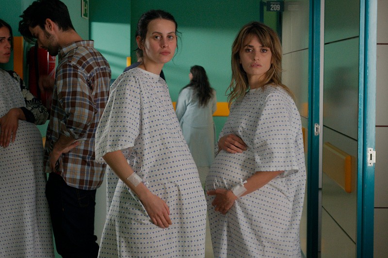 Dwie kobiety w ciąży w szpitalnych koszulach stoją na jasno oświetlonym korytarzu szpitalnym, patrząc w kamerę. W tle widać inne osoby, w tym mężczyznę i kobietę.