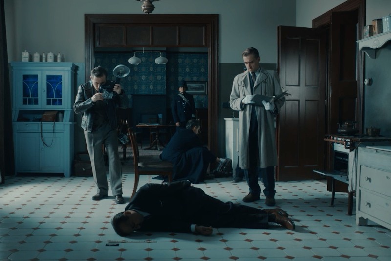 Miejsce zbrodni w pokoju w stylu vintage przedstawia mężczyznę leżącego twarzą do dołu na kafelkowej podłodze, otoczonego przez śledczych — jeden robi zdjęcia, drugi pisze notatki, a trzeci bada teren. W tle widać policjanta.