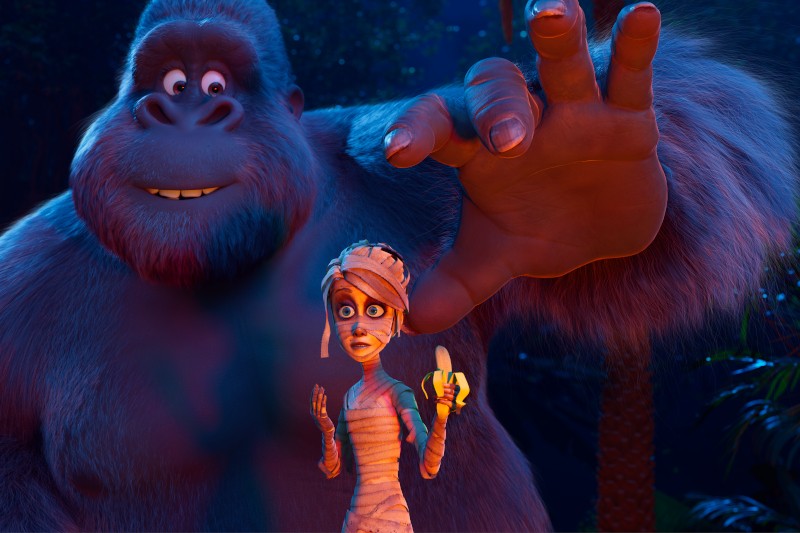 Duży, przyjaźnie wyglądający animowany goryl wyciąga rękę w kierunku zaskoczonej mumii trzymającej banana w ciemnym, przypominającym dżunglę otoczeniu.