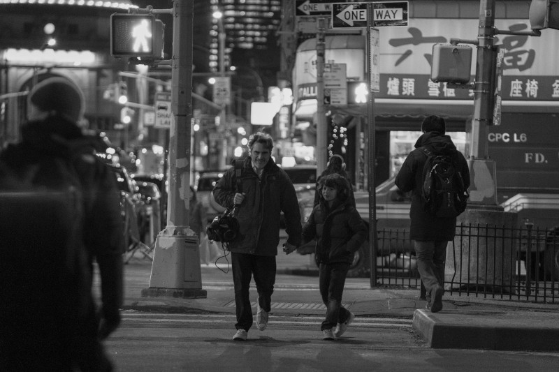 Na czarno-białym zdjęciu widać mężczyznę i dziecko trzymające się za ręce, przemierzających ruchliwą ulicę miasta nocą, w otoczeniu pieszych i podświetlonych znaków.
