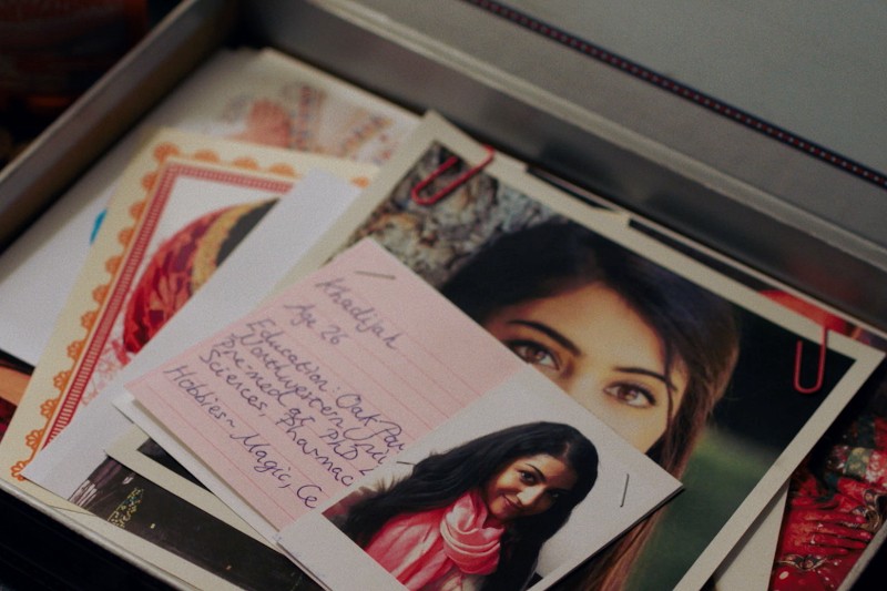 Zbliżenie otwartego pudełka zawierającego odręcznie pisane notatki, zdjęcia uśmiechniętej kobiety o ciemnych włosach i listy, niektóre spięte czerwonym spinaczem. Widoczna notatka jest napisana na różowym papierze.