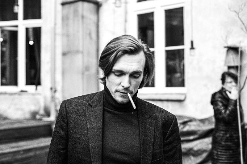 Mężczyzna o średniej długości włosach, ubrany w ciemny garnitur i golf, patrzy w dół z papierosem w ustach. Inna osoba stoi rozmazana w tle w pobliżu budynku z dużymi oknami. Zdjęcie jest czarno-białe.