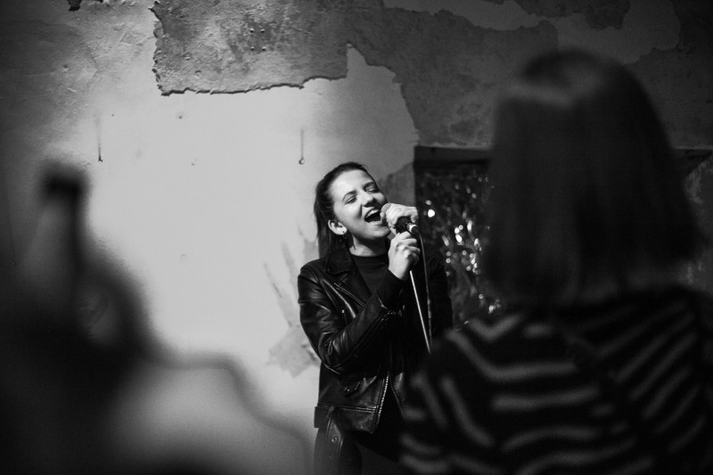 Kobieta w skórzanej kurtce z pasją śpiewa do mikrofonu na scenie. Na pierwszym planie widać niewyraźne sylwetki widzów, a za nią widać teksturowaną, zniszczoną ścianę.