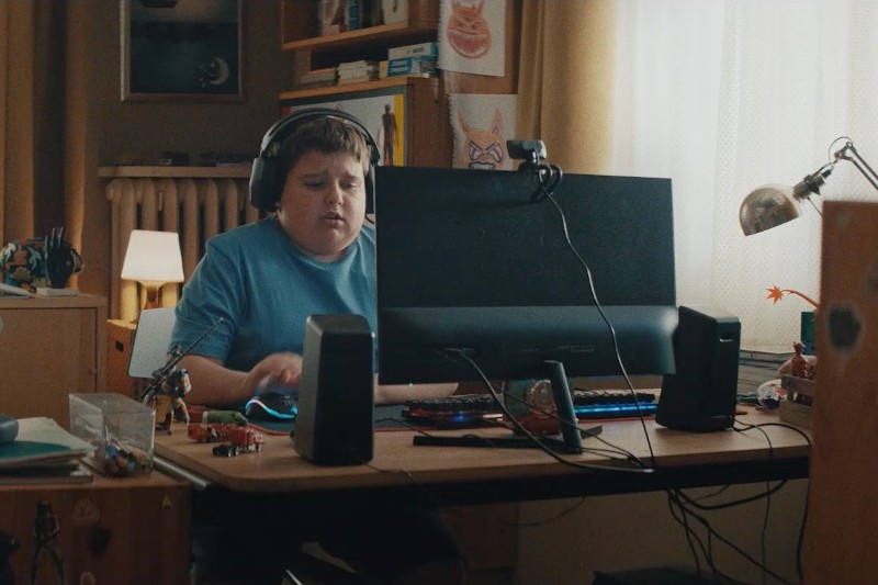 Chłopiec w słuchawkach siedzi przy biurku w przytulnym pokoju, skupiony na monitorze komputera z klawiaturą i myszką. Jego biurko jest zagracone zabawkami i gadżetami, a światło dzienne wpada przez zasłoniętą zasłoną szybę.