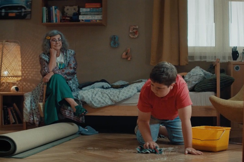 Chłopiec w czerwonej koszuli klęczy na podłodze, szorując szmatką obok żółtego wiadra, podczas gdy starsza kobieta z siwymi włosami i okularami siedzi na łóżku, patrząc na niego i uśmiechając się. Pokój wygląda przytulnie i zamieszkany.