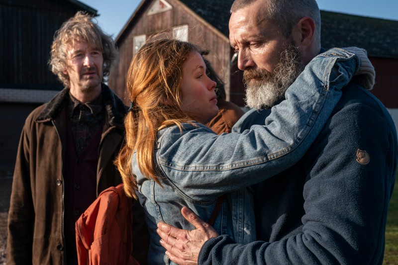 Kobieta z rudymi włosami i dżinsową kurtką obejmuje brodatego mężczyznę na zewnątrz, oboje wyglądają na wzruszonych. Inny mężczyzna z kręconymi włosami i brązowym płaszczem stoi w tle i patrzy na nich. Za nimi widać stodoły.