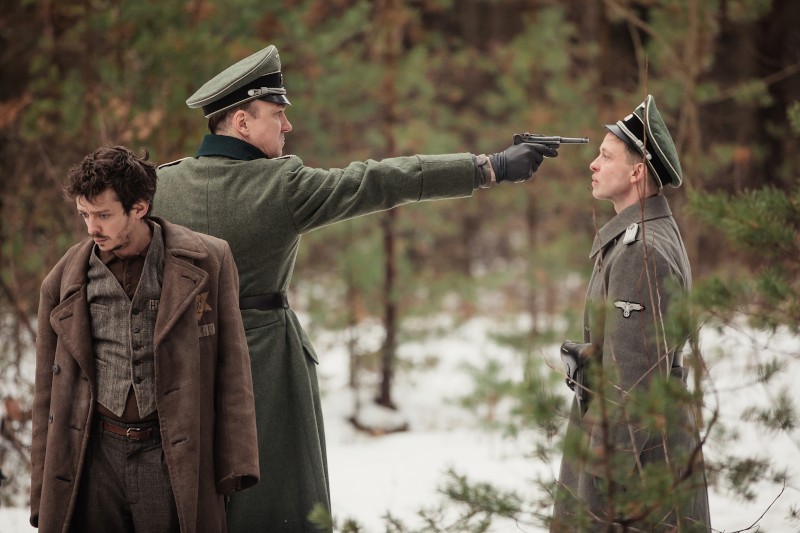 Mężczyzna w mundurze nazistowskim celuje pistoletem w głowę innego mężczyzny w mundurze, podczas gdy trzeci mężczyzna w cywilnym ubraniu z żółtą gwiazdą odwraca wzrok. Stoją w zaśnieżonym lesie.