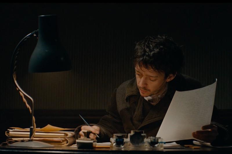 Mężczyzna siedzi przy biurku pod czarną lampą, trzymając kartkę papieru i pisząc długopisem. Na biurku w słabo oświetlonym pokoju znajdują się stosy dokumentów i butelek z atramentem.