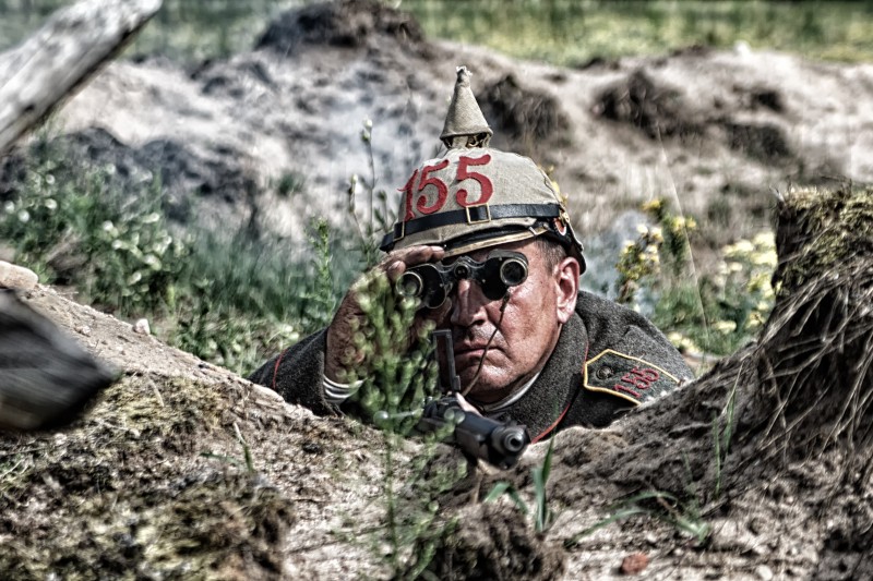 Żołnierz w historycznym mundurze wojskowym i hełmie oznaczonym numerem 155 kuca w okopie, patrząc przez lornetkę z karabinem przed sobą. Trawa i ziemia otaczają żołnierza na zewnątrz.