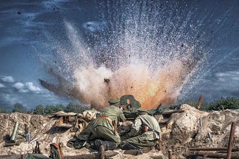 Dwóch żołnierzy w mundurach wojskowych kryje się w okopie, gdy przed nimi dochodzi do potężnej eksplozji, w wyniku której w powietrze, pod dramatycznym, zachmurzonym niebem, wzbijają się odłamki i dym.