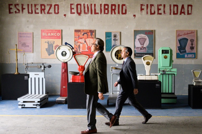 Dwóch mężczyzn w garniturach przechodzi obok wystawy starych wag i kolorowych plakatów na ścianie. Słowa Esfuerzo, Equilibrio, Fidelidad są namalowane nad wystawą pogrubionymi czerwonymi literami.