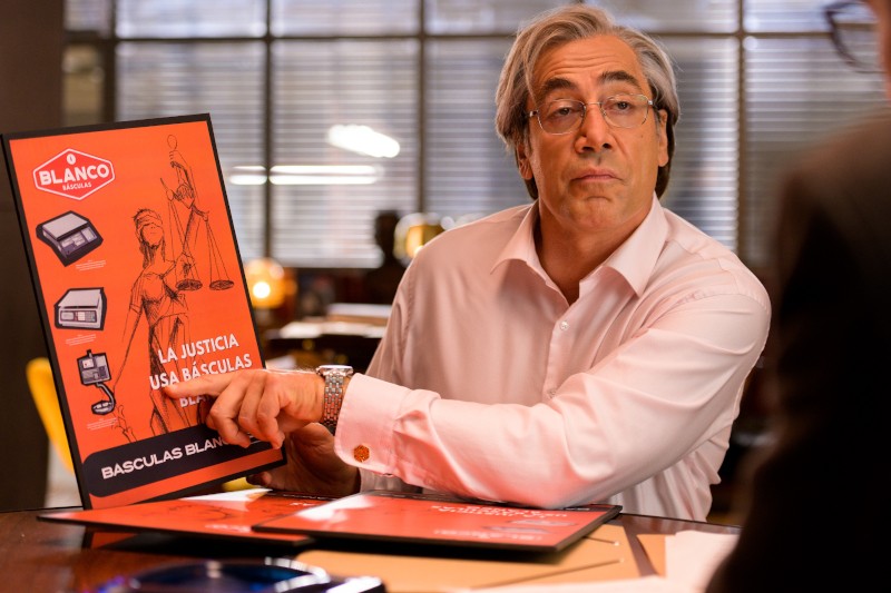 Mężczyzna o siwych włosach i okularach siedzi przy biurku i wskazuje na jaskrawopomarańczowy plakat reklamowy Básculas Blanco, na którym znajduje się waga i napis La Justicia Usa Básculas w języku hiszpańskim.