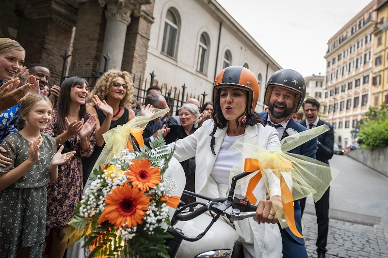 Panna młoda i pan młody w kaskach jadą na udekorowanym kwiatami i wstążkami skuterze, otoczeni wiwatującymi i oklaskującymi gośćmi, na ulicy miasta.
